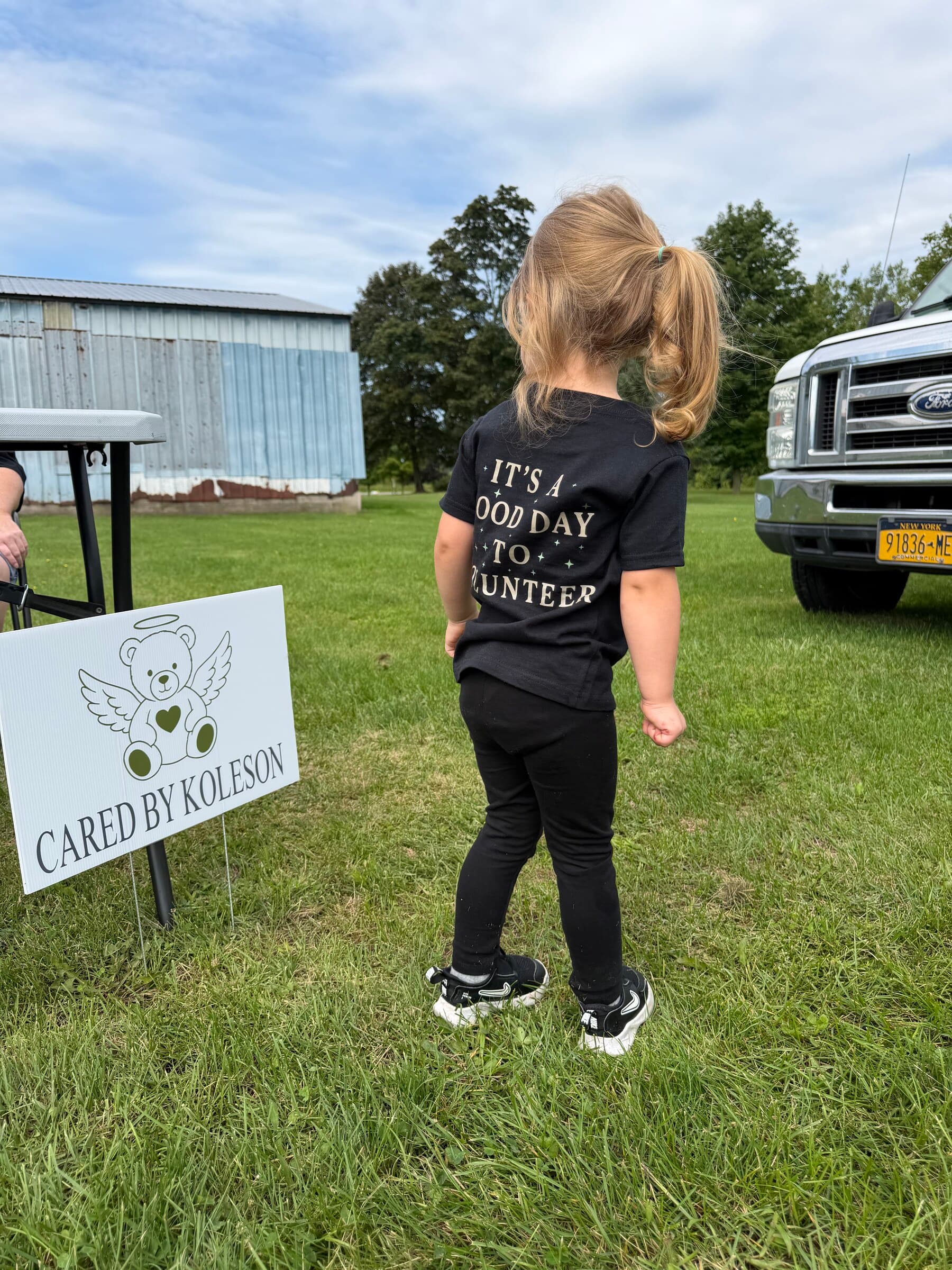 A young volunteer beside the Cared By Koleson yard sign, wearing an 'It's a good day to volunteer' tee